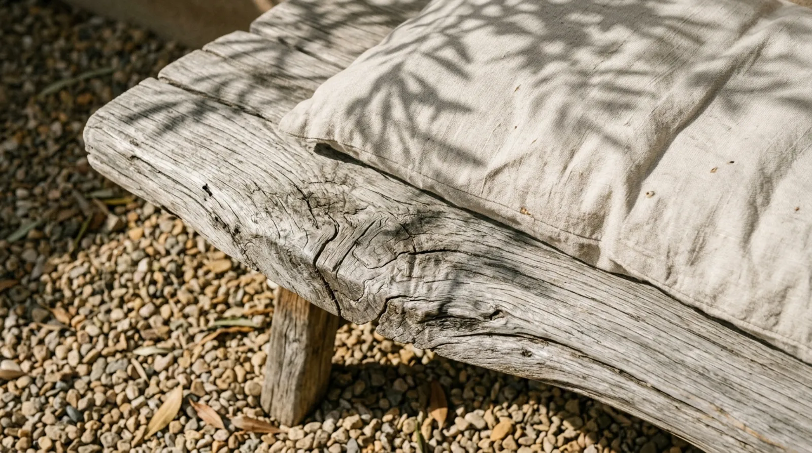 Weathered olive-wood bench with a linen cushion and leaf shadows.